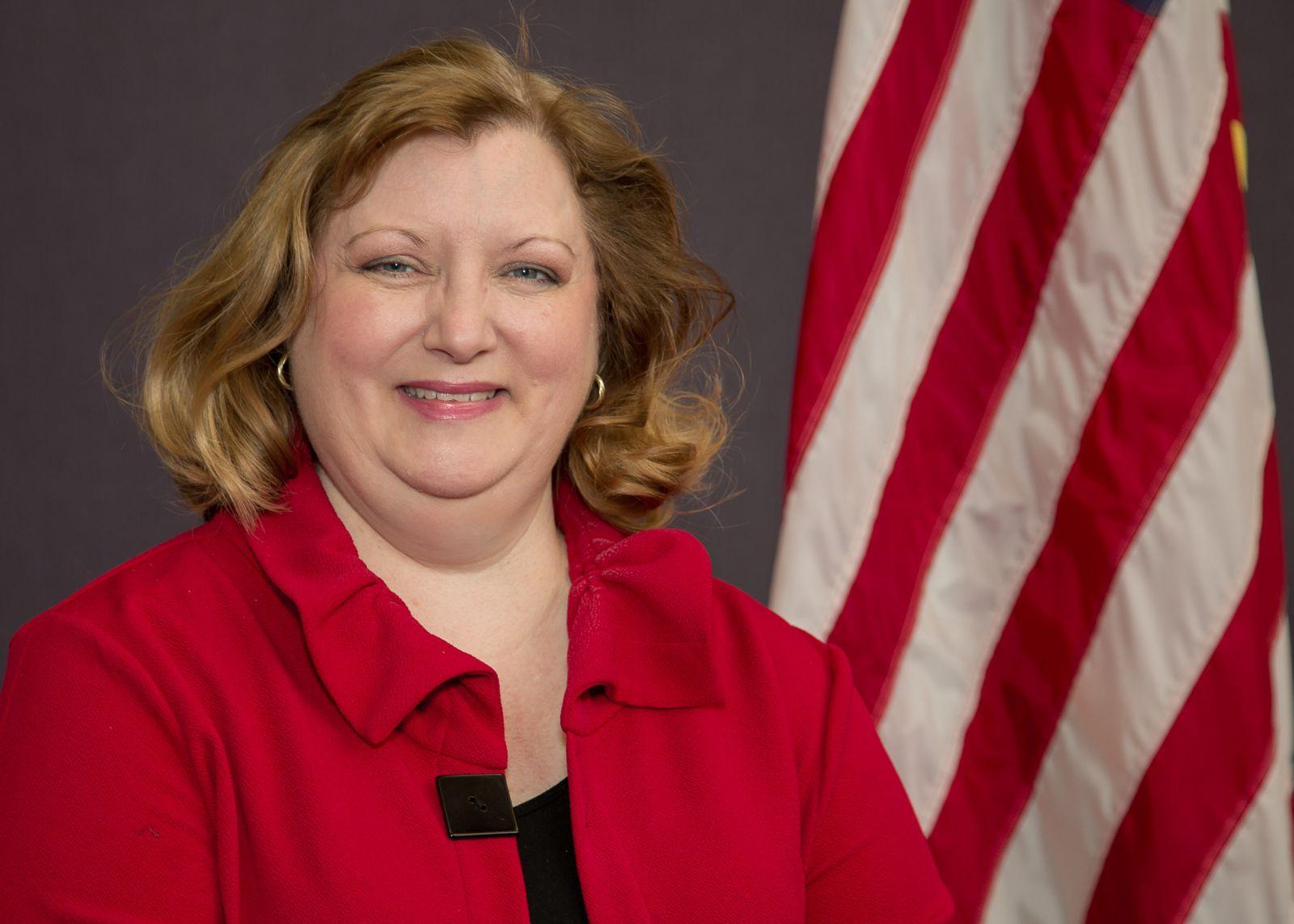 A blonde, white caucasian, Gena Sapp, is wearing a bright red collared jacket over a black shirt and is pictured with the United States flag standing in the back left against a grey background.